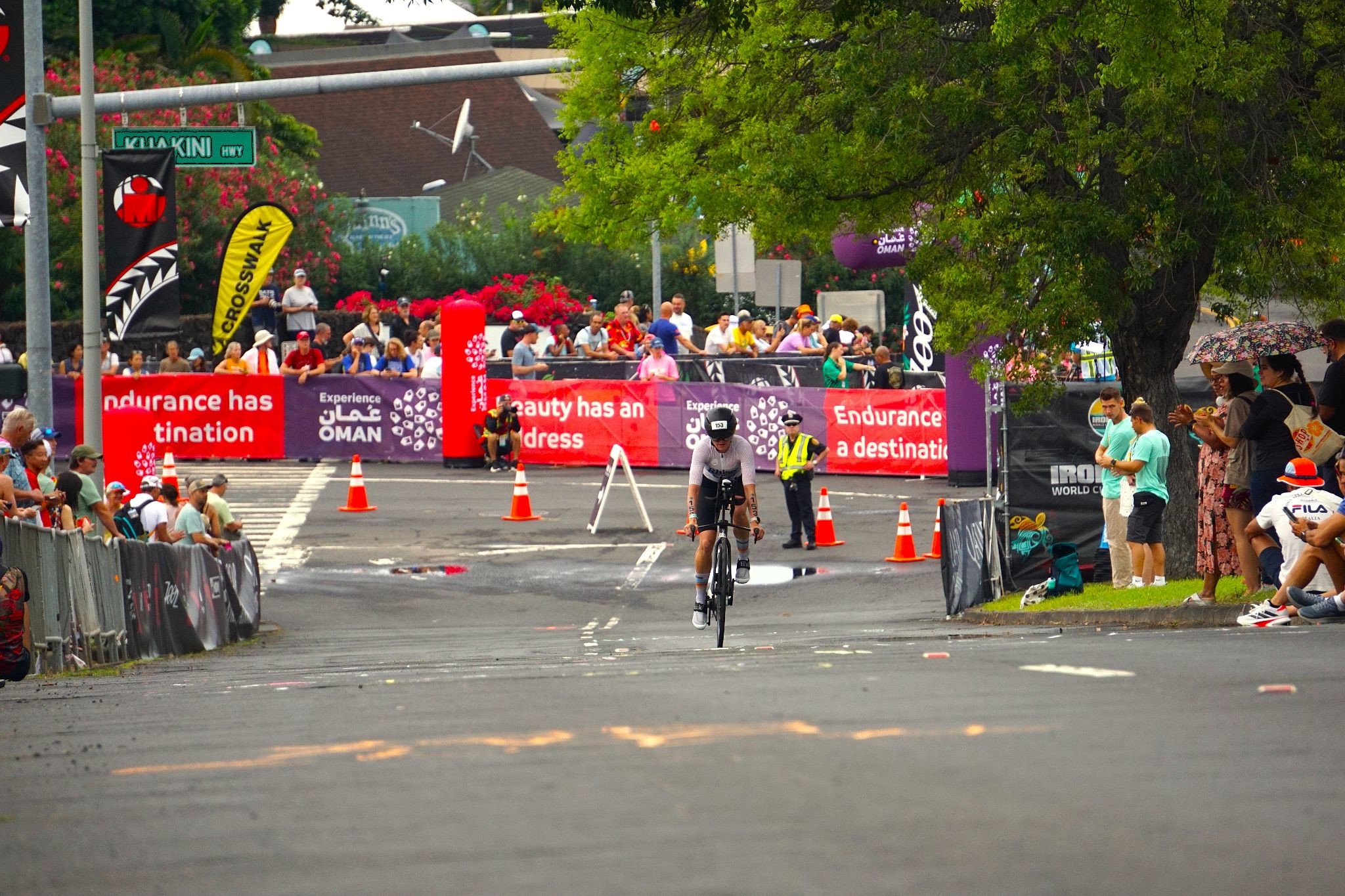 A lone cyclist pushes through the final stretch of the bike course, demonstrating the dedication, the struggle, and the ultimate triumph of the IRONMAN race. Surrounded by cheering spectators and race banners, this image captures the atmosphere as athletes near the end of the windswept 112-mile ride on the Queen Ka'ahumanu Highway.